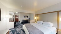 Located at 1383 Washington Ave in Albany, NY, this thoughtfully designed hotel room offers a comfortable and functional layout across a single floor. The spacious main area is anchored by a plush king-size bed set within a defined sleeping nook, framed by a warm wood headboard and complemented by elegant globe wall sconces. A large window floods the sleeping area with natural light, creating a bright and welcoming ambiance. Bold, patterned carpeting in deep navy and grey tones adds a contemporary flair throughout.

A wall-mounted flat-screen TV and a coordinating wood media console provide entertainment, while a plush armchair offers a relaxed seating option. The room transitions seamlessly into a dedicated workspace featuring a modern desk, ergonomic chair, and a tall floor lamp—ideal for productivity. A compact kitchenette, equipped with a microwave, mini-refrigerator, and sink set into dark cabinetry with a wood countertop, adds everyday convenience.

The private bathroom is finished with large-format white tile flooring and a striking navy herringbone accent wall, creating a bold yet clean aesthetic. A floating wood vanity with an integrated sink, a large mirror, and a shower/tub combination complete the well-appointed space.