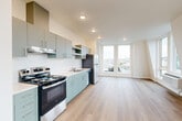 Located at 401 Union Ave SE in Olympia, WA, this modern one-bedroom, one-bathroom unit offers a thoughtfully designed open-concept layout finished with light hardwood-style flooring throughout. The combined kitchen and living area forms the heart of the unit, featuring a well-appointed kitchen with two-tone cabinetry — sage green lower cabinets paired with gray upper cabinets — stainless steel appliances including a refrigerator, dishwasher, and electric range, recessed lighting, and a clean white countertop. Large corner windows in the living area flood the space with natural light and offer pleasant views of the surrounding Olympia cityscape and greenery. The bedroom is a bright, comfortable retreat with oversized windows on two walls that continue the theme of abundant natural light and outdoor views, complemented by a spacious double sliding-door closet. A hallway connects the main living space to the bedroom, bathroom, and an additional closet. The combined bathroom and laundry room features a stacked washer and dryer unit, a soaking tub, and sage green vanity cabinetry with a white countertop, maintaining the cohesive modern aesthetic found throughout the unit. Clean white walls and minimalist finishes give the entire space a fresh, contemporary feel.