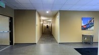 This single-story commercial space is defined by a long, expansive corridor that serves as the central artery of the building. The hallway features warm beige and tan-painted walls that create an inviting, neutral atmosphere, complemented by a drop tile ceiling fitted with recessed lighting that evenly illuminates the length of the passageway. The flooring transitions from large-format tile at the entrance to a patterned, multi-toned carpet runner that lines the corridor, adding texture and visual interest underfoot. Dark-trimmed doorways are positioned along both sides of the hallway, providing access to individual rooms or suites, while wall-mounted light fixtures offer additional ambient lighting between doorways. The overall design is clean and functional, with a professional aesthetic suited for a hospitality or office environment. The repetition of architectural elements — from the ceiling grid to the evenly spaced doors — creates a sense of depth and order, making the space feel organized and purposeful. The building's layout and finishes reflect a well-maintained, move-in-ready interior designed for high-traffic use.