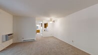 This single-floor residential unit offers a straightforward, functional layout designed for comfortable everyday living. The main living area is generously proportioned, featuring neutral wall-to-wall carpeting, crisp white walls, and a ceiling fan with light fixture that adds a touch of practicality. A baseboard heating system and wall-mounted AC unit ensure year-round climate comfort. The living space opens toward the kitchen through a pass-through counter, creating a connected and convenient flow between areas.

The kitchen is well-equipped with warm wood-toned cabinetry extending to the ceiling for ample storage, white appliances including a range, dishwasher, and refrigerator, a double-basin sink, and light tile flooring illuminated by a skylight-style ceiling panel.

The bedrooms are carpeted and bright, with windows that frame views of lush greenery outside. Each room includes closet storage with louvered sliding doors, and baseboard heaters run along the walls. One bedroom features convenient direct access to the kitchen area.

The bathroom is finished in a clean, neutral palette with tile flooring, a wood-toned vanity with mirror, and a full-size soaking tub with shower combination. The overall ambiance of the unit is light, airy, and move-in ready.