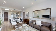 This thoughtfully designed single-floor space showcases a seamless open-concept layout that effortlessly blends the kitchen, dining, and living areas into one cohesive and inviting environment. The interior is finished in a clean, neutral palette of whites and warm grays, complemented by recessed lighting and smooth ceilings that enhance the sense of airiness throughout.

The kitchen is appointed with stainless steel appliances, warm wood cabinetry, dark countertops, and a breakfast bar with white counter stools — ideal for casual dining or entertaining. Pendant lighting adds a refined touch above the bar area.

The dining area features a sleek glass-top table surrounded by modern white chairs, creating an elegant yet approachable setting. A rich wood-toned sideboard serves as a stylish transitional piece between the dining and living spaces.

The living area is anchored by a plush gray sofa adorned with soft accent pillows, paired with a glass coffee table and a cozy area rug. A large decorative mirror with a wood frame reflects light across the room, while wall art and curated décor add personality. The warm hardwood-style flooring flows continuously throughout, tying all areas together with warmth and sophistication.