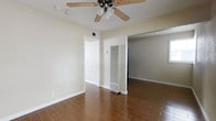 This single-floor space presents a straightforward and functional layout designed for comfortable everyday living. The main living area is finished with warm wood-look tile flooring laid in a classic brick pattern, adding texture and visual warmth to the room. Neutral beige walls provide a bright, clean backdrop throughout, complemented by crisp white baseboards and door trim. A ceiling fan with a light fixture is centrally mounted, offering both ventilation and ambient lighting. A wall-mounted HVAC unit provides climate control directly within the living space. The room opens through a wide doorway into an adjacent space, suggesting a connected and open flow between areas. A secondary doorway leads to what appears to be a hallway or additional room, enhancing the sense of accessibility and circulation throughout the floor plan. Ceiling-mounted vents indicate a supplemental HVAC system as well. The overall ambiance is simple, neutral, and move-in ready, with a no-frills aesthetic that provides a versatile canvas for personalization. The single-floor design ensures ease of movement throughout the entire space without the need for stairs.