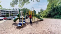 This outdoor playground offers a well-equipped recreational space designed for active play. The centerpiece is a multi-station play structure finished in bold green, yellow, and blue, featuring two slides — including a tunnel slide and an open slide — along with a climbing ladder, staircase entry with handrails, and interactive activity panels that encourage learning and engagement. The structure is thoughtfully designed to accommodate children of varying ages and skill levels. The ground surface beneath and around the play equipment is covered with wood chip mulch, providing a cushioned, safety-conscious base. The playground is framed by a lush canopy of mature deciduous trees, offering natural shade during warmer months and creating a pleasant, park-like atmosphere. The surrounding landscape includes dense greenery and open sky, contributing to an inviting outdoor environment. A residential building is visible in the background, suggesting this playground may serve as an amenity for a nearby community. The space is open and accessible, with ample room around the equipment for supervision and free movement.