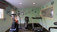 Located at 1900 N US-17 Hwy in Mount Pleasant, South Carolina, this single-floor fitness facility offers a practical and welcoming environment for exercise. The room is finished with light mint-green walls adorned with colorful circular floral decals, giving the space an upbeat and energetic atmosphere. Recessed ceiling lighting ensures the area is well-lit, while windows allow for natural light to filter in, enhancing the overall brightness of the room. The flooring features rubber matting, providing a safe and durable surface suited for fitness activities. The facility is equipped with essential cardio machines, including a treadmill, an upright stationary bike, and an elliptical or stair-climbing machine, offering users a variety of cardiovascular workout options. Wall-mounted accessories, including a sanitizing station, reflect attention to cleanliness and user convenience. The compact yet functional layout makes efficient use of the available space, ensuring all equipment is accessible. Overall, the facility presents a clean, cheerful, and purpose-driven environment well-suited for residents or visitors looking to maintain their fitness routine.