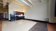 Located at 2715 Barber Ave in Cleveland's Ohio City neighborhood, this single-floor unit showcases a compelling blend of industrial character and modern updates. The open-plan layout maximizes the generous sense of space, with soaring ceilings that expose white-painted ductwork and conduit overhead, lending the interior an authentic loft aesthetic.

The main living area features a striking combination of polished terrazzo flooring and reclaimed brick-tone flooring, creating distinct visual zones without the use of walls. Large steel-framed windows along one wall allow natural light to pour in while offering views of the surrounding neighborhood, complemented by dark curtain panels for privacy.

The kitchen is positioned along one wall and is outfitted with rich dark-wood cabinetry extending nearly to the ceiling, dark granite countertops, and a full suite of black appliances including a refrigerator, range, and over-the-range microwave. Track and cove lighting illuminate the kitchen area effectively.

The bathroom maintains the same warm design sensibility, featuring dark-wood vanity cabinetry with a white countertop and sink, brushed nickel fixtures, a full tub-shower combination, and brick-tone tile flooring that ties the space together cohesively. The overall ambiance is urban, functional, and characterized by a balance of raw industrial details and thoughtful finishes.