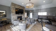 This single-story space exudes a refined yet comfortable ambiance, centered around a dramatic floor-to-ceiling stacked stone accent wall that serves as the room's focal point. A gas fireplace is built into the stone wall, adding warmth and character, while a large flat-screen television is mounted above it, making the space both functional and inviting. The seating arrangement features a collection of tufted leather club chairs and a sofa in a rich charcoal tone, gathered around a rustic round coffee table atop a classic patterned area rug — creating an intimate and cohesive conversation area. An elegant drum chandelier with candelabra-style lighting hangs from the vaulted ceiling, casting a warm glow throughout the room. The walls are finished in a cool gray tone, complemented by horizontal shiplap paneling details that add texture and visual interest. Wood-look plank flooring runs throughout, grounding the space with a warm, natural element. Natural light filters in through multiple windows, further brightening the interior. The overall design balances rustic and contemporary elements, resulting in a welcoming, well-appointed communal lounge.
