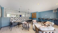 Located at 4673 Lake Valley Drive in Lisle, Illinois, this single-floor community clubhouse serves as a welcoming social hub for Lakeside Apartments. The lounge is defined by its bold teal-painted walls, which create a vibrant and contemporary atmosphere throughout the space. Light-toned ceramic tile flooring runs continuously across the entire area, contributing to a clean and open feel.

The seating area is thoughtfully arranged with a mix of upholstered lounge chairs and ottomans in neutral gray tones, accented with colorful throw pillows. Café-style round tables paired with sleek chairs provide casual dining or gathering spots, while counter-height bar seating along the wall offers an alternative workspace or social area with a stone-topped surface.

A distinctive design feature is the large mirrored partition with a bold black-and-white geometric patterned tile backsplash, which visually expands the space and adds a dynamic graphic element. Recessed ceiling lighting and pendant fixtures provide layered illumination throughout. Decorative woven wall art on the teal accent walls adds warmth and texture.

Large glass sliding doors and full-length windows at the rear of the space flood the lounge with natural light and offer direct views and access to the outdoor swimming pool area, seamlessly connecting the interior to outdoor amenity spaces. A branded entry mat at the front entrance completes the polished, community-focused design.