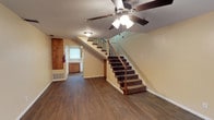 This two-story home presents a straightforward and functional layout designed for comfortable everyday living. The main level features rich, dark hardwood-style flooring that extends throughout the open living area, creating a warm and cohesive foundation for the space. A ceiling fan with integrated lighting is centrally positioned overhead, providing both airflow and ambient illumination.

A carpeted staircase with a classic wood handrail and decorative iron balusters serves as the central architectural feature of the main level, connecting the two floors in a visually prominent and practical way. The warm yellow-toned walls lend a welcoming atmosphere throughout.

Toward the rear of the main floor, a glimpse into the kitchen area reveals wood cabinetry and a window that allows natural light to filter into the space. A floor vent indicates a central HVAC system for climate control, and a fire extinguisher is conveniently mounted near the kitchen entry, reflecting attention to safety.

The area beneath the staircase is thoughtfully utilized, adding to the overall sense of efficient use of space. Clean white baseboards trim the perimeter of the room, providing a crisp contrast to the darker flooring. The second floor, accessible via the staircase, expands the living space further, offering additional rooms above the main level.