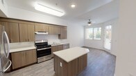 This single-floor space showcases a thoughtfully designed open-concept layout where the kitchen and living area flow seamlessly together. The kitchen is outfitted with warm wood-toned cabinetry that extends to the upper walls, providing generous storage. A central island with a clean white countertop serves as both a workspace and a natural divider between the kitchen and living area. Stainless steel appliances, including a gas range and dishwasher, add a modern, functional touch, complemented by an under-cabinet range hood and a stainless steel sink. The flooring throughout features wood-look planks in a light grey tone, contributing to the contemporary aesthetic. The adjoining living area is bright and open, illuminated by natural light streaming through large windows dressed with blinds, as well as a sliding door that leads to the outdoors. A ceiling fan with a light fixture provides both comfort and additional illumination. The white walls and clean lines throughout create a fresh, neutral palette that is versatile and inviting.
