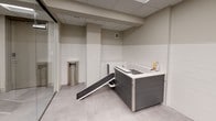 This single-floor amenity suite offers two distinct functional areas designed to serve residents and their pets. The shared laundry room is accessed through a glass-paneled door and houses multiple top-load and front-load washing machines along with dryers, all finished in white and arranged along the walls. The room features a drop ceiling with recessed panel lighting, smooth neutral-toned walls, and light-colored concrete-style flooring, creating a clean and utilitarian environment.

Adjacent to the laundry area is a thoughtfully designed pet spa, enclosed with floor-to-ceiling glass partitions for visibility and easy supervision. The space is decorated with framed photographic prints of dogs, adding a welcoming character. A purpose-built grooming tub with a built-in entry ramp accommodates pets of varying sizes, while grooming supplies are conveniently stored nearby. White subway tile wainscoting lines the lower half of the walls, complementing the neutral upper wall tones. Stainless steel waste receptacles are positioned throughout both areas. The suite maintains a cohesive, modern aesthetic with consistent flooring and ceiling treatments, ensuring a practical yet polished environment.