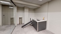 This single-floor amenity suite offers two distinct functional areas designed to serve residents and their pets. The shared laundry room is accessed through a glass-paneled door and houses multiple top-load and front-load washing machines along with dryers, all finished in white and arranged along the walls. The room features a drop ceiling with recessed panel lighting, smooth neutral-toned walls, and light-colored concrete-style flooring, creating a clean and utilitarian environment.

Adjacent to the laundry area is a thoughtfully designed pet spa, enclosed with floor-to-ceiling glass partitions for visibility and easy supervision. The space is decorated with framed photographic prints of dogs, adding a welcoming character. A purpose-built grooming tub with a built-in entry ramp accommodates pets of varying sizes, while grooming supplies are conveniently stored nearby. White subway tile wainscoting lines the lower half of the walls, complementing the neutral upper wall tones. Stainless steel waste receptacles are positioned throughout both areas. The suite maintains a cohesive, modern aesthetic with consistent flooring and ceiling treatments, ensuring a practical yet polished environment.