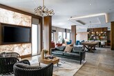 Located at 5 Freer Street in Lynbrook, New York, this thoughtfully designed single-floor amenity space offers a dynamic blend of comfort, style, and functionality. The open-plan layout flows seamlessly across distinct zones, each curated with high-end finishes and contemporary furnishings.

The main lounge area anchors the space with rich leather seating, a striking onyx-framed entertainment wall with a flat-screen TV, and an artful area rug. Expansive floor-to-ceiling windows frame outdoor courtyard views and flood the interior with abundant natural light. Copper and globe pendant lighting add warmth and visual interest throughout.

A dedicated sitting nook features plush teal velvet armchairs arranged around a fireplace, creating an intimate retreat within the larger open space. Nearby, a games area accommodates a full-size pool table and shuffleboard, both set beneath sleek pendant lights against textured accent walls.

The private dining area showcases an oval walnut table surrounded by upholstered chairs with brass-tipped legs, beneath mirrored globe pendants and a bold teal accent wall with a mounted display screen — ideal for gatherings or events.

The wet bar is outfitted with rich walnut cabinetry, marble countertops, a built-in microwave, and under-counter refrigeration, providing convenient service for the entire lounge. Herringbone-patterned hardwood flooring and stone tile unite the zones, while warm wood columns and designer lighting complete the refined, modern aesthetic.