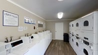 This shared laundry facility is designed for practicality and efficiency, offering residents a clean and organized space to handle their laundering needs. The room features a generous row of white front-loading washing machines lined along one wall, each equipped with individual control panels for ease of use. Opposite, a bank of stacked dryer units maximizes the use of vertical space, providing ample drying capacity. The flooring throughout is a durable hardwood-style material in a warm wood tone, lending a polished and well-maintained appearance. Neutral taupe-toned walls and a smooth white ceiling with flush-mount lighting create a bright and welcoming atmosphere. Informational notices and instructional signage are posted on the walls, ensuring users have easy access to usage guidelines and relevant notices. A utility panel is also conveniently located within the room. A white interior door provides access to an adjoining area, and a trash receptacle is positioned nearby for added convenience. Crown molding along the ceiling adds a subtle architectural detail, elevating the overall finish of the space. The layout is thoughtfully organized to allow comfortable movement throughout the room.