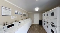 This shared laundry facility is designed for practicality and efficiency, offering residents a clean and organized space to handle their laundering needs. The room features a generous row of white front-loading washing machines lined along one wall, each equipped with individual control panels for ease of use. Opposite, a bank of stacked dryer units maximizes the use of vertical space, providing ample drying capacity. The flooring throughout is a durable hardwood-style material in a warm wood tone, lending a polished and well-maintained appearance. Neutral taupe-toned walls and a smooth white ceiling with flush-mount lighting create a bright and welcoming atmosphere. Informational notices and instructional signage are posted on the walls, ensuring users have easy access to usage guidelines and relevant notices. A utility panel is also conveniently located within the room. A white interior door provides access to an adjoining area, and a trash receptacle is positioned nearby for added convenience. Crown molding along the ceiling adds a subtle architectural detail, elevating the overall finish of the space. The layout is thoughtfully organized to allow comfortable movement throughout the room.