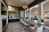 Located at 23400 Kingsland Blvd in Houston, TX, this sophisticated living space offers a refined and contemporary atmosphere designed for comfort and elegance. The single-floor layout centers around a well-appointed lounge area anchored by a sleek, linear fireplace set into a striking stone accent wall, above which a flat-screen television is mounted. Soaring ceiling heights amplify the sense of openness, while a multi-pendant chandelier with cylindrical glass tubes serves as a dramatic focal point overhead. The seating arrangement features a plush gray sofa complemented by coordinating accent chairs adorned with bold geometric throw pillows, all arranged around a mirrored glass coffee table atop a richly textured area rug. Architectural columns clad in natural stone define the space and extend into an adjoining area, where warm amber wall sconces and circular ceiling fixtures maintain a cohesive, upscale ambiance. A built-in refreshment station with dark cabinetry, marble-style countertops, and a coffee machine lines one wall beneath decorative shelving and patterned wallcovering. Large windows dressed with floor-to-ceiling gray drapes invite natural light while offering views of the surrounding area. The overall aesthetic blends modern luxury with warm, inviting tones throughout.