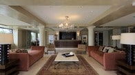 This single-floor lobby presents a sophisticated and welcoming atmosphere defined by thoughtful architectural and design elements. At its center hangs an ornate crystal chandelier that serves as the focal point of the space, casting warm ambient light across the cream-toned ceiling fitted with recessed lighting. Classical white columns flank the open floor plan, adding a sense of formality and structure to the layout.

The reception area is anchored by a sleek, dark wood desk positioned against a deep-toned accent wall, flanked by wall sconces and framed artwork that contribute to the refined aesthetic. The seating arrangement in the foreground features two plush, rose-toned sofas and a pair of woven accent chairs, all centered around a rectangular white-topped coffee table resting on a richly patterned Persian-style area rug. The light marble tile flooring ties the elements together, enhancing the sense of openness throughout. Large windows or glass doors visible at the periphery suggest natural light and outdoor views, contributing to the bright and airy quality of the overall space.