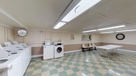 The Village Park laundry facility is a generously sized, single-floor communal space designed to serve residents with efficiency and convenience. The room is finished with a classic green-and-cream checkered vinyl tile floor that lends a clean, timeless aesthetic throughout the space. Bright fluorescent lighting is installed across the ceiling, ensuring the entire area is well-lit for practical use.

The facility is outfitted with a substantial number of machines, including both top-loading and front-loading washers as well as stacked and standalone dryers, providing residents with ample options to accommodate varying laundry needs. A stainless steel utility table offers a dedicated workspace for sorting and preparing laundry, while a large folding table provides additional surface area for organizing clean clothes.

A row of waiting chairs lines one wall, offering a comfortable place for residents to wait during their laundry cycles. A wall-mounted mirror, a clock, and posted laundry guidelines contribute to the room's functional and organized character. Crown molding details along the upper walls add a subtle architectural touch.

A windowed exterior door at one end of the facility allows for natural light to filter in and provides direct access to the outdoors. The overall layout of the space is practical and well-organized, making it a reliable and accessible amenity for all Village Park residents.