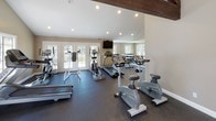 This single-level fitness center is defined by its striking architectural character, most notably the vaulted ceiling with prominent exposed dark wood beam trusses that add visual warmth and a sense of height to the space. The layout is divided into two connected zones, together offering a comprehensive workout environment.

The cardio area is well-appointed with multiple treadmills, elliptical trainers, stationary bikes, and a stair climber, all arranged to maximize use of the floor space. Dark rubber flooring throughout provides durability and cushioning underfoot. Recessed lighting supplements the generous natural light that streams in through several windows and sets of glass French doors, which also provide direct outdoor access and views of the surrounding landscape.

The strength training zone features a multi-function cable and weight machine alongside open floor space for stretching or functional exercise. Warm beige walls and white trim maintain a clean, neutral aesthetic throughout. A wall-mounted television in the cardio area and a dumbbell rack along the mirrored wall round out the amenities, making this fitness center a fully functional and thoughtfully designed space for varied workout routines.
