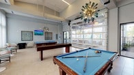 This contemporary game and social lounge is designed for recreation and relaxed socializing, offering a well-appointed single-floor layout filled with natural light and modern design elements. At the heart of the space are multiple full-size billiard tables with blue felt surfaces, positioned across light-toned tile flooring that runs throughout the room. Dramatic sputnik-style chandeliers with teal glass accents hang from the soaring ceilings, providing both ambient illumination and a striking visual focal point.

A dedicated lounge area features rounded upholstered chairs, a compact arcade-style game console, and wall-mounted flat-screen TVs, making it an ideal spot for spectators and casual conversation. Colorful abstract artwork adorns the walls, adding a curated, gallery-like quality to the interior.

Along one wall, a bank of floor-to-ceiling windows dressed in sheer curtains floods a separate seating nook with soft, diffused daylight. Pedestal café tables paired with mint-toned barrel chairs complete this bright corner, offering a quieter retreat within the space.

One of the most distinctive architectural features is the large glass panel garage door that opens directly onto an outdoor covered terrace, seamlessly connecting the interior to an al fresco lounge area. This indoor-outdoor connectivity enhances the airy, resort-like ambiance that defines the space.