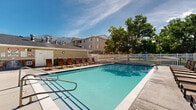 Located in Pleasant Grove, Utah, this well-appointed outdoor amenity area offers a resort-style experience against a stunning backdrop of the Wasatch Mountains. The centerpiece is a generously sized swimming pool with crystal-clear blue water, surrounded by an expansive concrete sun deck lined with rows of lounge chairs — ideal for relaxing and soaking up Utah's abundant sunshine. Adjacent to the pool, a bubbling hot tub provides a dedicated space for relaxation, finished with classic blue tile accents and accessible via built-in steps. A separate shaded patio area features wrought-iron tables and chairs sheltered beneath large umbrellas, offering a comfortable spot for outdoor dining or socializing. The entire complex is enclosed by a crisp white picket fence, lending the space a clean, welcoming aesthetic while maintaining privacy. A single-story amenity building sits along the perimeter, providing convenient access to restrooms and other facilities. Mature trees are thoughtfully positioned throughout the area, offering natural shade and greenery. The open sky above and panoramic mountain views beyond the fence create a scenic, airy atmosphere that defines this well-maintained outdoor gathering space.
