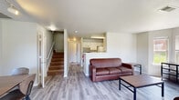 Located at 3535 E Tenth St in Greenville, NC, this two-story unit offers a well-organized layout across two levels. The main floor features an open-concept living area with light hardwood-style flooring, neutral walls, and large windows that invite abundant natural light and views of the surrounding neighborhood. A bay-style window arrangement in the living room enhances the sense of space, while a front seating area with direct access to the outdoors adds convenience and connectivity to the exterior.

The kitchen is positioned adjacent to the living area in a half-wall, pass-through configuration, fitted with white cabinetry, a full-size refrigerator, an electric range, and a sink — providing efficient functionality. A stacked washer and dryer unit is conveniently integrated within the space, offering in-unit laundry.

Upstairs, the bedrooms feature plush carpeting, natural window light with views of greenery, and practical furnishings including desks, dressers, and closets with paneled sliding doors — well-suited for everyday living. A bathroom on the upper level includes a single vanity with a wide mirror and hardwood-style flooring. The overall design is clean and functional, with a neutral color palette maintained throughout both floors.