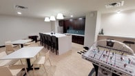 This single-floor amenity space is thoughtfully designed to serve as a social hub, combining a functional kitchenette, comfortable dining areas, and recreational entertainment in one open layout. The space features light wood-tone flooring throughout, contributing to a clean and welcoming atmosphere.

The kitchenette anchors one end of the room, outfitted with rich dark espresso cabinetry, a stainless steel refrigerator, and a teal herringbone tile backsplash that adds a subtle pop of color. Ample counter space and upper and lower cabinets provide practical storage and prep areas for gatherings.

A large white-topped central island serves as the focal point of the space, surrounded by dark upholstered bar-height chairs, accommodating multiple guests comfortably. Pendant lights hang above the island, providing warm, focused illumination. Additional dining tables with lighter chairs are positioned nearby, offering flexible seating arrangements.

Recessed lighting throughout the ceiling ensures the entire space is well-lit. A tabletop hockey game is visible in the foreground, highlighting the recreational character of the room. The overall design balances utility with a relaxed, community-oriented ambiance.