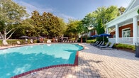 Located at 100 Ivey Park Ln in Norcross, Georgia, this outdoor amenity space centers around a generously sized freeform swimming pool with clear turquoise water. The pool is framed by a classic brick coping edge and surrounded by an expansive paver deck finished in a cobblestone-style pattern, providing ample space for relaxation and foot traffic alike. Rows of sleek lounge chairs line the poolside, several shaded by navy blue market umbrellas, offering comfortable spots to unwind in the Georgia sun. A pergola structure is visible toward the back of the deck, adding a defined architectural focal point to the outdoor setting. The pool area is enclosed by a wrought iron fence, ensuring a secure and well-defined boundary. Mature trees — including magnolias and crepe myrtles — surround the perimeter, creating a lush, canopied atmosphere that provides natural shade and privacy. A classic red brick clubhouse with a covered porch and white railings serves as a prominent backdrop, lending a warm, traditional Southern character to the space. The combination of manicured landscaping, thoughtful seating arrangements, and charming architectural details makes this outdoor pool area an inviting and well-appointed community amenity.