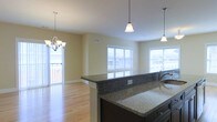 This single-floor space showcases an open-concept layout that seamlessly connects the kitchen and living areas, creating a spacious and functional environment. The kitchen is a standout feature, equipped with dark-toned cabinetry and rich granite countertops that extend across both the main workspace and a peninsula-style island, providing ample prep space and a natural division between the kitchen and the adjoining living area. Pendant lighting hangs above the island, complementing a elegant chandelier that anchors the living space, together adding warmth and visual interest to the interior. The flooring throughout is light-toned hardwood, lending a clean, cohesive look that enhances the sense of openness. Natural light is a defining characteristic of this space, with multiple large windows along the exterior walls allowing sunlight to pour in from several directions. The neutral, warm-toned walls keep the interior feeling bright and inviting. Ceiling-mounted HVAC vents indicate a central air system, adding to the practicality of the space. Overall, the combination of quality finishes, thoughtful lighting, and an airy layout gives this space a polished and welcoming ambiance.