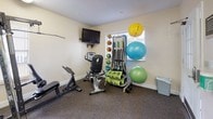 This compact yet comprehensively outfitted fitness center is designed to support a wide variety of workout routines. The space is single-level and features durable, rubber-textured flooring suited for high-traffic exercise use, with light-toned walls and bright overhead fluorescent lighting that create a clean and energizing atmosphere.

The cardio area is equipped with a treadmill, an elliptical trainer, and an upright stationary bike, offering multiple options for cardiovascular exercise. A large wall-mounted mirror spans one side of the room, visually expanding the space and allowing users to monitor their form during workouts. A wall-mounted flat-screen television adds entertainment value for those longer cardio sessions.

For strength training, the facility includes a multi-function cable and weight machine with various attachment points, as well as a weight bench for free-weight exercises. A dedicated accessory station provides an impressive array of functional fitness tools, including medicine balls in multiple sizes, resistance bands, foam rollers, and stability balls in assorted sizes — all neatly organized on a rolling storage rack.

Entry to the fitness center is through a white-framed exterior door with glass paneling, offering a glimpse of the outdoor surroundings, as well as interior French doors connecting to an adjacent space. Posted fitness room rules near the entrance underscore the facility's community-oriented nature.