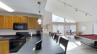 Located at 1101 Sutters Mill Lane in Knoxville, Tennessee, this single-floor communal space is thoughtfully designed to accommodate both everyday use and group gatherings. The open-concept layout seamlessly connects a fully equipped kitchen with a spacious dining and lounge area, creating a functional and welcoming environment throughout.

The kitchen features warm oak cabinetry, black appliances including a range and over-the-range microwave, and a generous dark-topped bar counter with seating along one side, illuminated by elegant pendant lights suspended from above. Track lighting along the ceiling further enhances the workspace and dining area.

Flowing naturally from the kitchen, the dining area is defined by striking vaulted ceilings that rise toward a dramatic bank of floor-to-ceiling angled windows. These expansive windows flood the space with natural light and offer views of the surrounding outdoor greenery, creating an airy, sunlit atmosphere. Light tile flooring throughout reflects the natural light and adds to the bright, open feel of the room. Several café-style bistro tables with wrought iron chairs are arranged near the windows, providing additional casual seating options. The overall ambiance is clean, functional, and inviting — well-suited for community dining, socializing, or everyday activities.