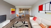 This well-appointed 1-bedroom, 1-bathroom unit offers a thoughtfully designed open-concept layout that seamlessly connects the living, dining, and kitchen areas. The living room features wood-look flooring, neutral walls, and a large window that invites natural light and outdoor views, creating a bright and welcoming atmosphere. The kitchen is outfitted with modern grey cabinetry with wood-tone upper panels, a white tile backsplash, under-cabinet lighting, and a full suite of stainless steel appliances including a refrigerator, range, and over-the-range microwave. A compact dining area with a round table and seating sits at the transition between the kitchen and living space, making the layout ideal for everyday living and entertaining. The bedroom is generously proportioned, featuring multiple windows with views to the outdoors, wood-look flooring, and a spacious built-in closet with bifold doors offering ample storage. The full bathroom is clean and functional, with a soaking tub and shower combo, a white vanity, and rich wood-tone flooring that adds warmth to the space. Throughout the unit, a consistent use of neutral tones, modern fixtures, and durable flooring creates a cohesive, contemporary aesthetic.