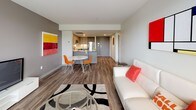 This well-appointed 1-bedroom, 1-bathroom unit offers a thoughtfully designed open-concept layout that seamlessly connects the living, dining, and kitchen areas. The living room features wood-look flooring, neutral walls, and a large window that invites natural light and outdoor views, creating a bright and welcoming atmosphere. The kitchen is outfitted with modern grey cabinetry with wood-tone upper panels, a white tile backsplash, under-cabinet lighting, and a full suite of stainless steel appliances including a refrigerator, range, and over-the-range microwave. A compact dining area with a round table and seating sits at the transition between the kitchen and living space, making the layout ideal for everyday living and entertaining. The bedroom is generously proportioned, featuring multiple windows with views to the outdoors, wood-look flooring, and a spacious built-in closet with bifold doors offering ample storage. The full bathroom is clean and functional, with a soaking tub and shower combo, a white vanity, and rich wood-tone flooring that adds warmth to the space. Throughout the unit, a consistent use of neutral tones, modern fixtures, and durable flooring creates a cohesive, contemporary aesthetic.
