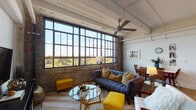 Located in Memphis, TN, this 2-bedroom, 2-bathroom unit blends industrial character with contemporary comfort across a single floor. The open-concept living and dining area is the heart of the space, anchored by a dramatic wall of floor-to-ceiling factory-style windows that flood the room with natural light and offer sweeping views of the surrounding treetops. Exposed brick walls, painted structural beams, visible ductwork, and soaring ceilings reinforce the building's industrial heritage, while a ceiling fan and polished concrete floors add functional appeal.

The living area features a tufted blue sofa, vibrant yellow accent ottomans, and a glass coffee table, creating a warm and inviting atmosphere. A dedicated dining nook with wicker chairs sits adjacent, benefiting from the same generous window light. The kitchen is fitted with white cabinetry, granite-style countertops, a double sink, and full appliances including a dishwasher.

The primary bedroom offers a clean, bright retreat with crisp white bedding and a clerestory window. The second bedroom retains the industrial aesthetic with an original concrete column as a standout architectural detail and accommodates two beds. Both bathrooms feature white cabinetry, granite countertops, and concrete flooring, with one including a full tub-shower combination.