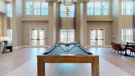 Located at Tapestry Park in Indian Land, SC, this sophisticated community clubhouse delivers an impressive blend of modern design and versatile amenity spaces across a generous single-floor layout. The space is defined by dramatically soaring double-height ceilings that amplify the sense of openness throughout, while large floor-to-ceiling windows and glass panel doors flood the interior with abundant natural light and provide views of the surrounding community.

The main gathering area centers around a full-size billiards table set beneath a striking rectangular pendant light fixture and an eye-catching sculptural chandelier, both of which complement the refined aesthetic. Light-toned large-format tile flooring flows seamlessly throughout, adding to the airy, contemporary feel.

A dedicated lounge zone features tufted upholstered seating, a built-in window bench, and an elegant marble-clad fireplace wall complete with a mounted flat-screen TV — creating an inviting focal point for relaxation. Adjacent to this is a well-appointed catering kitchen outfitted with dual-toned cabinetry, quartz countertops, a sink, appliances including a wine cooler, and its own large display screen. A dining area with pendant lighting and brick accent wall rounds out the space, making this clubhouse ideal for social gatherings and community events.