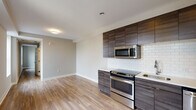 This single-floor unit showcases a clean, modern design aesthetic carried consistently throughout its layout. The kitchen serves as a focal point, equipped with a full suite of stainless steel appliances including a range with oven, built-in microwave, dishwasher, and refrigerator. Dark wood-grain cabinetry extends from counter height to the ceiling, providing generous storage, while white subway tile backsplash offers a crisp contrast. Light-toned countertops and a undermount sink complete the functional workspace.

The open layout allows the kitchen to flow naturally into the living area, which is finished with light hardwood flooring that extends throughout the unit, creating a cohesive and airy feel. The living space is well-proportioned, with neutral warm-gray walls and clean white baseboards contributing to a bright, contemporary atmosphere.

The bedroom is a calm, open room with the same hardwood flooring and neutral wall tones, offering a versatile blank canvas. The bathroom is finished to a modern standard, featuring a dark wood vanity with a white quartz-style countertop, an undermount sink, a large frameless mirror, and coordinating hardwood-style flooring, maintaining the unit's consistent design language.
