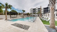 This expansive outdoor amenity area is designed with a resort-inspired aesthetic, centered around a large, shimmering swimming pool that serves as the focal point of the space. Tall palm trees are strategically placed throughout the courtyard, adding a tropical ambiance and natural shade. The pool deck is finished with light-toned stone pavers and features decorative rock garden accents, lending a clean and sophisticated look to the surroundings.

Lounge areas are thoughtfully arranged along the pool's perimeter, with chaise lounges, market-style string lights, and hammocks positioned on vibrant artificial turf lawns that provide comfortable relaxation zones. A dedicated putting green is tucked along one side of the complex, offering a recreational amenity for golf enthusiasts.

Covered walkways lined with white arched columns and pilasters extend along the building's ground level, providing shaded outdoor seating areas furnished with neutral-toned sofas, armchairs, and wicker accent tables. Ceiling fans installed beneath these colonnades enhance comfort during warmer months.

The surrounding multi-story building features a contemporary coastal architectural style, with white stucco facades, dark-trimmed balconies, and arched entryways finished with bold navy blue doors. The overall atmosphere is upscale, relaxed, and well-suited for outdoor leisure and community gatherings.