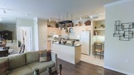 This single-floor residence showcases an open-concept layout that connects the kitchen, dining, and living areas into one cohesive, functional space. The kitchen serves as the heart of the home, featuring light cream cabinetry that extends to the ceiling, providing generous storage throughout. A central island with a dark countertop offers additional workspace and doubles as a casual gathering spot. A standout overhead pot rack hangs above the island, adding a practical and visually interesting element to the space. White appliances, including a refrigerator and microwave, complement the kitchen's clean, neutral palette. Track lighting runs across the ceiling, ensuring the entire area is well-illuminated. Adjacent to the kitchen, a built-in shelving nook with warm lighting creates a charming display and dining alcove, complete with a wooden chair and table. The flooring transitions from light tile in the kitchen area to rich dark hardwood in the living zone, clearly defining each functional space. The living area is visible in the foreground, furnished with a comfortable sofa and accented with candle holders. Crown molding adds a refined architectural detail throughout, contributing to the overall polished and welcoming ambiance of this thoughtfully designed home.