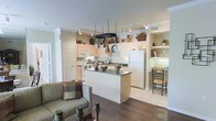 This single-floor residence showcases an open-concept layout that connects the kitchen, dining, and living areas into one cohesive, functional space. The kitchen serves as the heart of the home, featuring light cream cabinetry that extends to the ceiling, providing generous storage throughout. A central island with a dark countertop offers additional workspace and doubles as a casual gathering spot. A standout overhead pot rack hangs above the island, adding a practical and visually interesting element to the space. White appliances, including a refrigerator and microwave, complement the kitchen's clean, neutral palette. Track lighting runs across the ceiling, ensuring the entire area is well-illuminated. Adjacent to the kitchen, a built-in shelving nook with warm lighting creates a charming display and dining alcove, complete with a wooden chair and table. The flooring transitions from light tile in the kitchen area to rich dark hardwood in the living zone, clearly defining each functional space. The living area is visible in the foreground, furnished with a comfortable sofa and accented with candle holders. Crown molding adds a refined architectural detail throughout, contributing to the overall polished and welcoming ambiance of this thoughtfully designed home.
