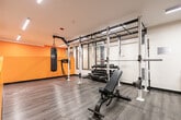 Located at 109 Vaughan Road in Toronto, Ontario, this fitness facility is a thoughtfully designed single-floor gym space that delivers a comprehensive workout environment. The interior is defined by a striking combination of bold orange accent walls and crisp white surfaces, creating an energetic and motivating atmosphere throughout.

The space is outfitted with a robust Dynamic power rack system, providing a central hub for strength training and functional fitness. A suspended heavy punching bag anchored to the rack adds a dedicated boxing and combat training element, complemented by a dumbbell rack offering a full range of free weights. Cardio equipment includes treadmills and elliptical cross-trainers, while a flat/incline adjustable bench and stability balls support versatile training routines.

Large floor-to-ceiling mirrors line the walls, visually expanding the space and allowing users to monitor their form during workouts. The flooring features durable wood-look planks that are both practical and aesthetically refined. Linear LED lighting is recessed into the smooth white ceiling, ensuring bright, even illumination throughout the entire floor. The open, single-room layout maximizes usable floor area, allowing for seamless movement between strength, cardio, and functional training zones — making this a highly functional and well-appointed fitness space.