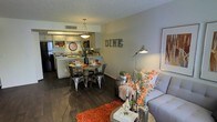 This single-floor residence offers an open-concept layout that integrates the living, dining, and kitchen zones into one cohesive, functional space. The living area is furnished with a light gray sofa accented by warm orange floral arrangements and a cozy throw, anchored by a woven area rug in a rich terracotta hue. A round dining table with industrial-style metal chairs occupies the central space, complemented by a statement pendant light overhead and a decorative "DINE" wall sign that adds a playful, personalized touch. An adjustable floor lamp provides additional ambient lighting to the living zone.

The kitchen, positioned at the rear of the open floor plan, features white cabinetry, a peninsula with bar-style seating, and stainless steel appliances including a refrigerator. The textured ceiling and recessed ventilation add to the functional design elements. Dark hardwood-style flooring flows consistently throughout, providing a rich contrast to the neutral wall tones. The overall ambiance is warm and inviting, combining contemporary furnishings with rustic and industrial accents for a well-balanced, lived-in aesthetic.