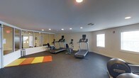 This fitness center is designed to support a well-rounded workout routine within a comfortable, single-floor layout. The space is outfitted with multiple treadmills and cardio machines, each equipped with individual display screens for tracking performance metrics. A dedicated stretching and floor exercise area is defined by brightly colored exercise mats, providing a functional zone for warm-ups, cool-downs, or bodyweight training. The room features recessed ceiling lighting that evenly illuminates the space, complemented by a natural light source from a window along one wall. A large wall-mounted mirror panel expands the sense of space while allowing users to monitor their form during workouts. The flooring consists of durable rubber gym matting, ideal for high-impact activity and equipment placement. Neutral-toned walls and a clean, uncluttered layout contribute to a focused and motivating exercise environment. The overall design prioritizes practicality and accessibility, making this fitness center a versatile facility suited for a range of fitness levels and training styles.