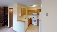 This two-floor residential space offers a practical and straightforward layout across its two levels. The kitchen is equipped with warm oak cabinetry, a double sink, a full suite of white appliances including a range and refrigerator, and light tile flooring. A breakfast bar peninsula provides a convenient transition between the kitchen and adjacent living areas. The living and dining areas feature a distinctive full-height exposed brick accent wall that adds character and texture to the space, complemented by light tile flooring on one level and plush carpeting on another, along with windows that bring in natural light and views of surrounding greenery. The bedrooms are carpeted and include louvered bi-fold closet doors, with windows providing additional natural light. The space includes multiple bathrooms: one features a vanity with oak cabinetry, a wall-mounted light bar, and a mirror, while another includes a full tub-shower combo with tile surround. Throughout the space, textured ceilings are consistent, and floor-plan views reveal a well-organized interior with a staircase connecting the two levels. Additional utility and storage areas with tile flooring round out the lower level, offering flexible functionality.