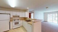 This single-floor residential unit is thoughtfully designed to offer a functional and comfortable living environment. The kitchen is well-appointed with white cabinetry, laminate countertops, and a full suite of white appliances including a refrigerator, electric range, dishwasher, and double-basin sink. A peninsula counter separates the kitchen from the living area, creating an open-concept layout that promotes easy movement and interaction between spaces. The living area is generously sized, featuring neutral carpet flooring, a ceiling fan with integrated light fixture, and baseboard heating. Warm wood-toned door trim and frames throughout the unit add a consistent design element that complements the light, neutral wall tones. The bedroom offers plush carpeting, a window with natural light, and baseboard heating, providing a quiet and comfortable retreat. The bathroom is notably designed with accessibility in mind, featuring multiple grab bars, a roll-in shower with handheld showerhead, tile flooring, and a vanity with sink. The overall layout, as seen in the dollhouse view, is compact yet efficiently organized, with a hallway connecting the main living areas to the private spaces. The unit presents a clean, move-in-ready appearance throughout.