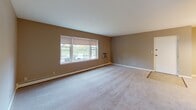 This single-floor residence offers a straightforward and functional layout, as revealed by the floor plan, which shows multiple rooms arranged around a central corridor. The living area is generously sized, finished with neutral-toned carpet and warm beige walls. A wide window along one wall allows natural light to fill the space while providing outdoor views, and a wall-mounted air conditioning unit and baseboard heating ensure year-round climate control. An adjoining dining area is distinguished by a classic chandelier overhead, adding a subtle decorative touch to the otherwise understated space.

The kitchen is equipped with honey-toned wood cabinetry both above and below the counter, a stainless steel sink, and a white electric range with overhead ventilation. Light-toned tile flooring ties the space together, giving it a clean and practical character.

The bedrooms continue the neutral carpet and beige wall palette, each featuring windows that bring in natural light and baseboard heating along the perimeter walls. The bathroom includes a full tub-shower combination with a white surround, a wood-framed vanity with a marble-style countertop, and warm terracotta mosaic tile flooring, completing the unit's consistent, functional aesthetic.