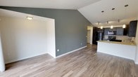 This single-floor space offers a thoughtfully designed open-concept layout that seamlessly connects the living area to the kitchen. The living room immediately impresses with its dramatic vaulted ceiling, which adds a strong sense of height and volume to the space. A bold slate-grey accent wall grounds the room and provides a striking contrast against the crisp white trim and ceilings. Light wood-look laminate flooring runs throughout, lending warmth and continuity to the open floor plan.

The kitchen is visible from the main living area and features dark cabinetry that creates a sleek, modern aesthetic. Stone-look countertops add texture and refinement to the workspace, while stainless steel appliances, including a full-sized refrigerator, contribute to the contemporary feel. Pendant lighting and a track lighting system above the kitchen ensure the space is both functional and well-lit.

A pass-through opening in the accent wall connects to an additional area, offering flexibility for use as a dining space or secondary living zone. The overall ambiance is clean, modern, and airy — a well-appointed space that balances bold design choices with practical, everyday livability.