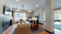 Carrick at Tonka Bay presents a refined single-floor community space designed with both comfort and functionality in mind. Rich hardwood flooring flows throughout, tying together each distinct area with warmth and cohesion. The main entrance welcomes residents and visitors through glass double doors, opening into a bright, open lobby accented by globe pendant lighting and a tray ceiling that adds architectural interest.

The central lounge is a standout gathering space, featuring a statement fireplace flanked by built-in shelving, a wall-mounted TV, and generous seating arranged beneath an iron hoop chandelier. Large windows flood the room with natural light and offer views of the surrounding community exterior. Adjacent to the lounge, a casual dining nook provides banquette-style seating with a nautical-inspired aesthetic, framed artwork, and more expansive windows.

The leasing office is enclosed by sleek black-framed glass walls, creating a professional yet transparent workspace stocked with property information and welcoming seating for consultations. A private conference room offers a more intimate setting, featuring a large white table, upholstered chairs, and a bold navy accent wall with curated artwork. Every corner of this space reflects a coastal-contemporary design sensibility with careful attention to detail.