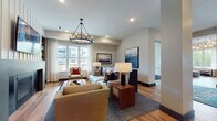 Carrick at Tonka Bay presents a refined single-floor community space designed with both comfort and functionality in mind. Rich hardwood flooring flows throughout, tying together each distinct area with warmth and cohesion. The main entrance welcomes residents and visitors through glass double doors, opening into a bright, open lobby accented by globe pendant lighting and a tray ceiling that adds architectural interest.

The central lounge is a standout gathering space, featuring a statement fireplace flanked by built-in shelving, a wall-mounted TV, and generous seating arranged beneath an iron hoop chandelier. Large windows flood the room with natural light and offer views of the surrounding community exterior. Adjacent to the lounge, a casual dining nook provides banquette-style seating with a nautical-inspired aesthetic, framed artwork, and more expansive windows.

The leasing office is enclosed by sleek black-framed glass walls, creating a professional yet transparent workspace stocked with property information and welcoming seating for consultations. A private conference room offers a more intimate setting, featuring a large white table, upholstered chairs, and a bold navy accent wall with curated artwork. Every corner of this space reflects a coastal-contemporary design sensibility with careful attention to detail.