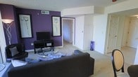 This single-floor space makes a strong visual statement through its thoughtful use of color and layout. The living area serves as the heart of the space, anchored by a deep purple accent wall that contrasts sharply against the surrounding white walls, creating a bold and modern aesthetic. Soft, neutral carpeting runs throughout, lending warmth underfoot and tying the rooms together cohesively.

The living room is comfortably arranged with a large, dark upholstered sofa facing a glass-top coffee table, which sits atop the carpeted floor. A flat-screen television is positioned on a low media console against the purple wall, beneath a framed botanical art piece that adds a decorative focal point. A tall, slender blue vase provides an additional accent that complements the room's color palette.

A textured ceiling adds subtle architectural interest overhead, while a ceiling vent indicates a central HVAC system throughout. The open doorway visible from the living area leads into an adjoining room, suggesting a well-connected and functional floor plan. The overall ambiance of the space is contemporary and curated, with intentional design choices that balance bold color with neutral tones to create an inviting, stylish interior.