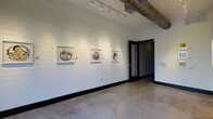 This single-floor gallery space offers a refined yet industrial aesthetic that creates an ideal setting for displaying artwork. The interior is defined by smooth white walls that serve as a neutral backdrop, currently showcasing a series of framed circular artworks mounted at eye level with accompanying labels. Polished concrete floors run throughout, lending the space a contemporary, urban character while remaining durable and easy to maintain.

The ceiling height is notably generous, contributing to an open, airy atmosphere. Exposed ductwork runs along the upper portion of the space, reinforcing the industrial design language. Adjustable track lighting is mounted along the ceiling, allowing for precise illumination of displayed works and flexible reconfiguration as exhibitions change.

Dark-framed doors with black hardware provide strong contrast against the white walls, adding to the space's modern sensibility. The dark baseboard trim throughout ties together the monochromatic palette. The overall layout is open and uncluttered, allowing visitors to move freely and engage with displayed pieces. The combination of clean lines, controlled lighting, and minimalist design creates a professional, gallery-quality environment well-suited for rotating art exhibitions.