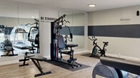Located at 18427 Studebaker Rd in Cerritos, CA, this fitness center is a well-appointed, single-room exercise space designed to support a full range of workout routines. The room features light wood-style flooring throughout, providing a clean and modern aesthetic that complements the neutral gray and white color scheme. A distinctive dark gray horizontal stripe runs along the walls, adding a bold design accent that ties the space together visually.

The cardio zone is equipped with multiple treadmills and an elliptical machine, all positioned along a wall lined with arched windows that invite natural light and offer views of the outdoors — an energizing backdrop for any workout. On the strength training side, the room includes a multi-function cable machine, an adjustable weight bench, and a spin bike, all placed atop protective rubber mats. Additional fitness accessories such as a plyo box, a step platform, and a stability ball are available for functional and cross-training exercises.

Large floor-to-ceiling mirrors along one wall visually expand the space and assist with form during workouts. Motivational wall decals — including "Be Stronger Than Your Excuses" and "-Motivation-" — reinforce an encouraging atmosphere. Recessed lighting ensures the room is well-lit at all hours. The space also includes a wall-mounted AC unit and sanitization stations near the entry for convenience and hygiene.